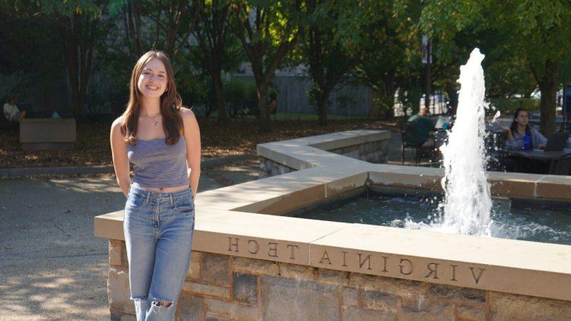 A steaming fountain in an enclosure marked Virginia Tech flows beside a young woman with long brown hair.